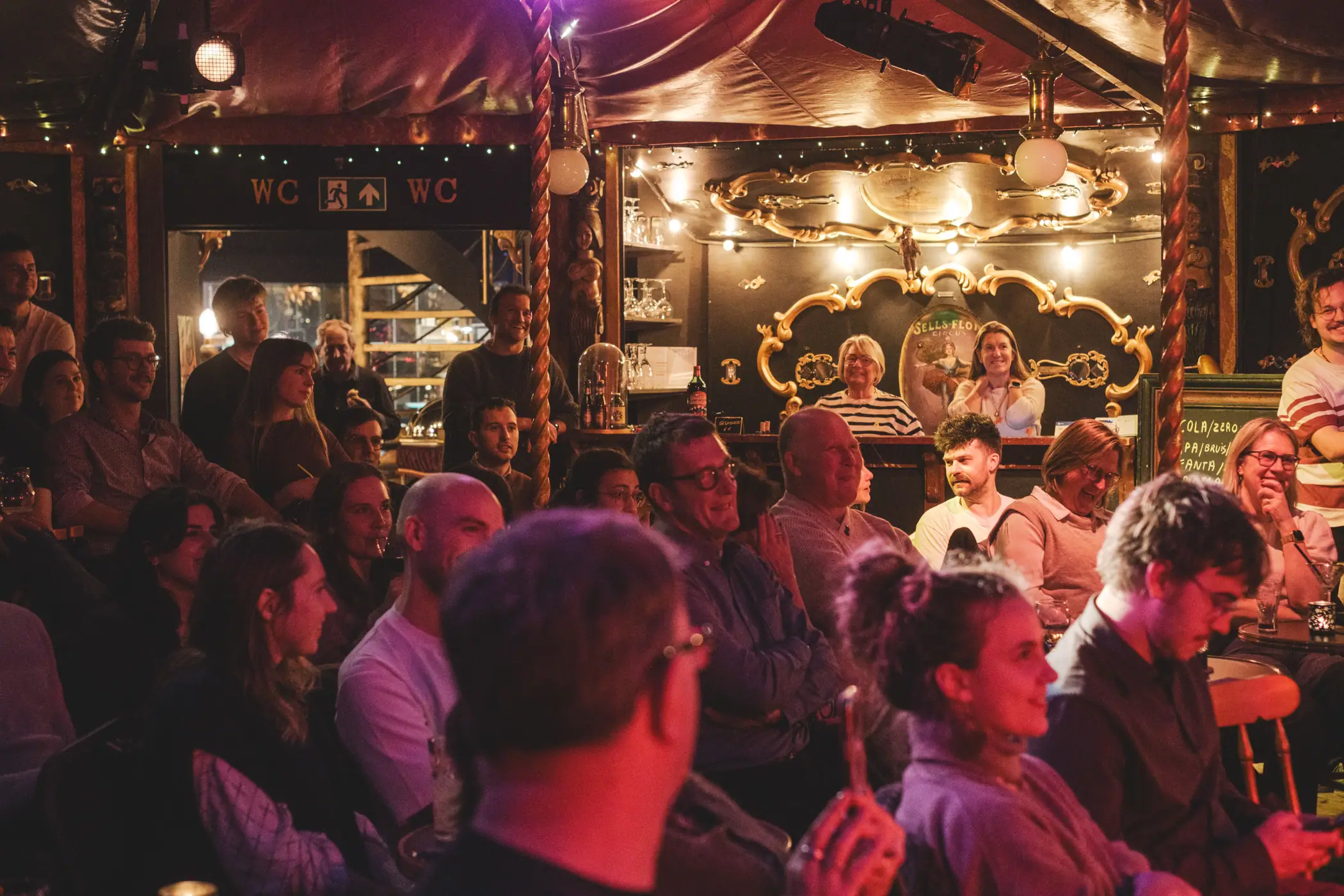 Volle zaal met warm licht: publiek van dichtbij, lachende gezichten, een bar met gouden lijsten op de achtergrond tijdens Echo Sessies
